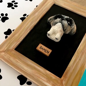 May include: A framed portrait of a dog with the name "Ramen" written on a brown plaque below the dog.