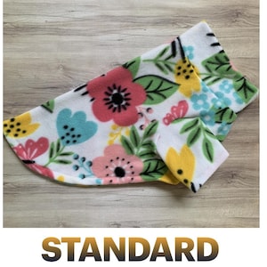 May include: A white fleece pet sweater featuring a colorful floral pattern with pink, yellow, and light blue flowers, along with green leaves and black dotted accents. The garment has a folded collar and a curved hemline for a comfortable fit.