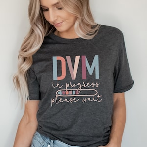 May include: A dark gray t-shirt with the text "DVM in progress please wait" in colorful letters. The text "in progress" is displayed as a progress bar with colorful blocks.