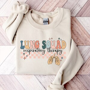 May include: A beige sweatshirt with the text "LUNG SQUAD respiratory therapy" in colorful letters. The text is surrounded by a checkered pattern and stars. A cartoon image of lungs is below the text.
