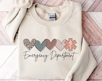 Può includere: Una felpa beige con una grafica di cinque cuori e un simbolo medico. I cuori sono di diversi colori e motivi, tra cui la stampa leopardata. Il testo "Emergency Department" è stampato sotto i cuori.
