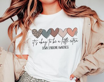 May include: White t-shirt with a line of colorful hearts and the text "it's okay to be a little extra" and "Down Syndrome Awareness".