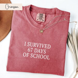 May include: Crimson-colored t-shirt with the text "I SURVIVED 67 DAYS OF SCHOOL" in white. The shirt has a crew neck and a small tag. A silver necklace with a pendant is draped across the shirt.