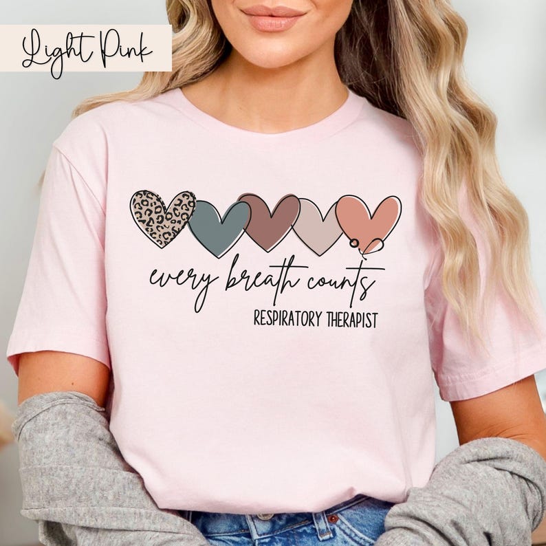 以下が含まれることがあります： 薄いピンクのTシャツに、異なる色と模様の5つのハートが並んでいます。ハートの下には「every breath counts」と「RESPIRATORY THERAPIST」の文字が印刷されています。