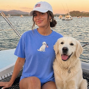 May include: A light blue t-shirt featuring an embroidered Labrador Retriever holding an American flag. The person is wearing a white baseball cap with an American flag design. A golden retriever dog is next to the person.
