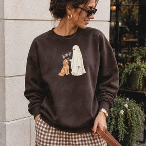 May include: A dark brown crewneck sweatshirt featuring a graphic print of a white ghost touching the nose of a golden-coloured dog. The text "BOO-P!" is printed in white above the dog. The sweatshirt has long sleeves and a ribbed neckline.