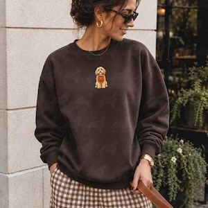 May include: A dark brown crewneck sweatshirt featuring a small, centered graphic of a light-colored puppy holding a small orange pumpkin. The fabric has a subtle, repeating tonal pattern of dog silhouettes across the entire garment.