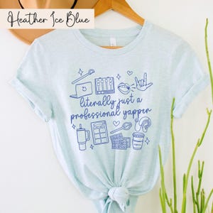 以下が含まれることがあります： 薄い青色のTシャツに、マイク、コーヒーカップ、ラップトップなどの青いグラフィックが描かれています。Tシャツには「literally just a professional yapper」という文字が印刷されています。