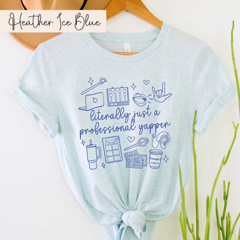 以下が含まれることがあります： 薄い青色のTシャツに、マイク、コーヒーカップ、ラップトップなどの青いグラフィックが描かれています。Tシャツには「literally just a professional yapper」という文字が印刷されています。