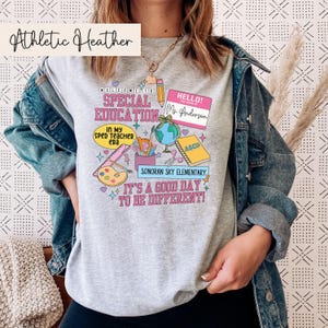 K&ouml;nnte beinhalten: Ein graues Sweatshirt mit einem bunten Design mit den Worten "Willkommen in der Sonderp&auml;dagogik" und "Es ist ein guter Tag, um anders zu sein!" Das Design enth&auml;lt auch einen Globus, einen Bleistift, ein Lineal, eine Farbpalette und ein Namensschild mit der Aufschrift "Hallo! Mein Name ist Frau Anderson".