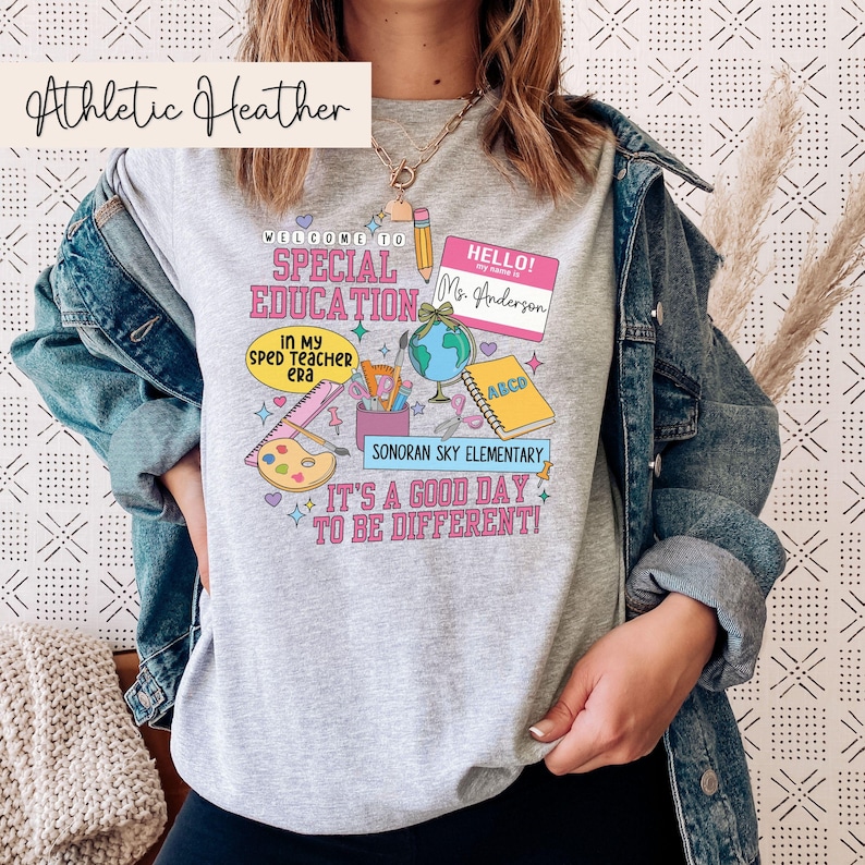 K&ouml;nnte beinhalten: Ein graues Sweatshirt mit einem bunten Design mit den Worten "Willkommen in der Sonderp&auml;dagogik" und "Es ist ein guter Tag, um anders zu sein!" Das Design enth&auml;lt auch einen Globus, einen Bleistift, ein Lineal, eine Farbpalette und ein Namensschild mit der Aufschrift "Hallo! Mein Name ist Frau Anderson".