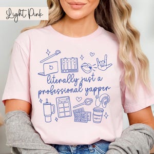 以下が含まれることがあります： 薄いピンクのTシャツに、青いグラフィックデザインが施されています。デザインには「文字通りプロのしゃべり屋」という文字と、ラップトップ、マイク、コーヒーカップ、ポッドキャストに関連するその他のアイコンが描かれています。