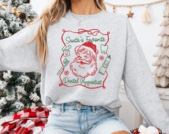 May include: A light grey sweatshirt with a red and green graphic that reads "Santa's Favorite Dental Hygienist." The graphic features Santa Claus, a toothbrush, toothpaste, and a tooth. The sweatshirt is a crew neck style.