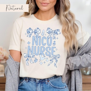 May include: A white t-shirt with a blue design that says "NICU NURSE" with blue bows, a baby bottle, a stork, and other baby-related icons.