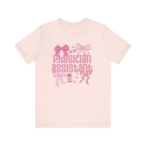 May include: Pink t-shirt with the text "Physician Assistant" in pink lettering. The text is surrounded by pink bows, pills, and a bandage.