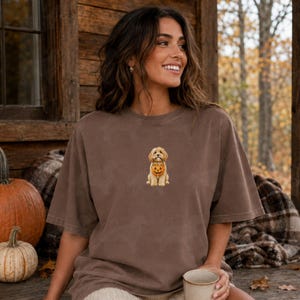 May include: A brown short-sleeved t-shirt featuring a small embroidered graphic of a golden-coloured dog sitting and holding a miniature orange jack-o'-lantern pumpkin bucket in its mouth. The shirt has a relaxed fit and a crew neckline.