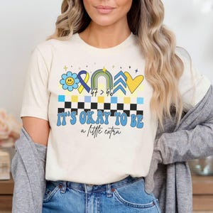 Pu&ograve; includere: Una maglietta bianca con un design blu e giallo che presenta un fiore, un nastro, un arcobaleno, una freccia e un cuore. Il testo "It's okay to be a little extra" &egrave; stampato sotto il design.