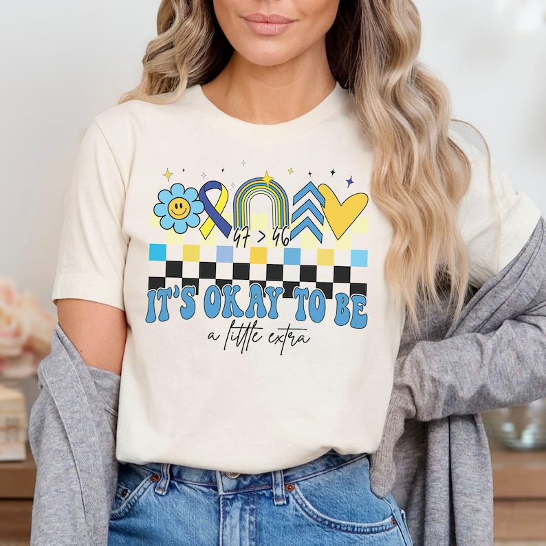 Pu&ograve; includere: Una maglietta bianca con un design blu e giallo che presenta un fiore, un nastro, un arcobaleno, una freccia e un cuore. Il testo "It's okay to be a little extra" &egrave; stampato sotto il design.