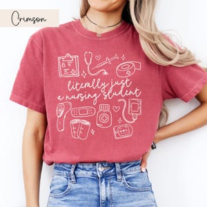 K&ouml;nnte beinhalten: Ein T-Shirt in Rotbraun mit einem wei&szlig;en Grafikdesign von medizinischen Utensilien und dem Text "literally just a nursing student".