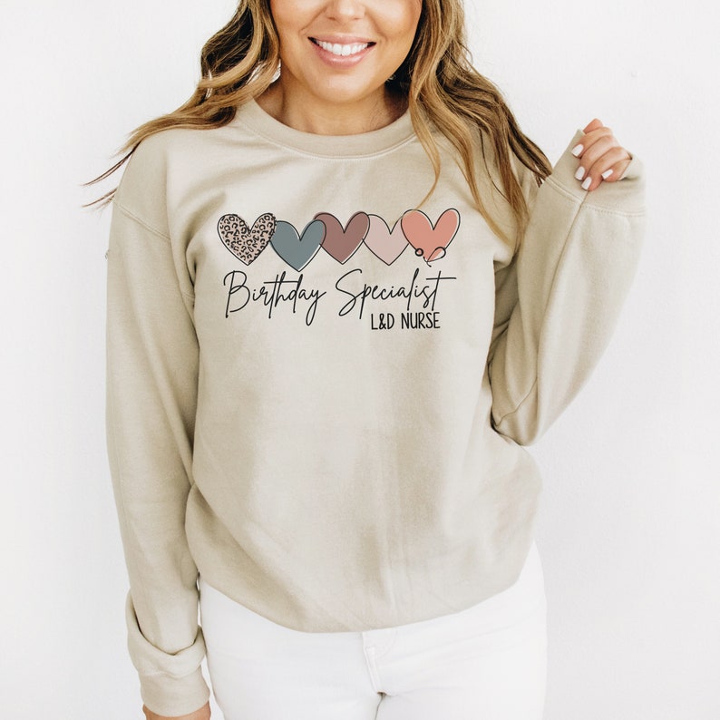 Op de afbeelding: Een beige sweatshirt met een afbeelding van vijf kleurrijke harten en de tekst "Verjaardagsspecialist L&D-verpleegkundige".