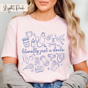 May include: Pale pink t-shirt featuring the phrase "literally just a doula" and blue line drawings of baby items, such as a nappy, bottle, and stork. The shirt is light pink.