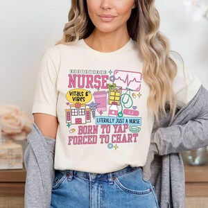May include: Cream-colored t-shirt with a colorful graphic design. The design includes the text "REGISTERED NURSE" and "VITALS & VIBES". Other text includes "LITERALLY JUST A NURSE", "BORN TO YAP", and "FORCED TO CHART". The graphic features medical-related icons.