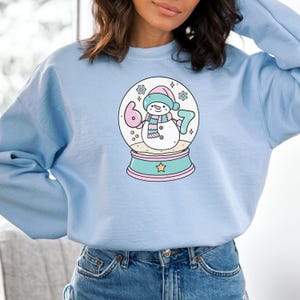 May include: Light blue sweatshirt featuring a snow globe design with a snowman wearing a pink hat and scarf. The snow globe has the numbers 6 and 7 on either side of the snowman. The base of the snow globe is teal with a pink trim.