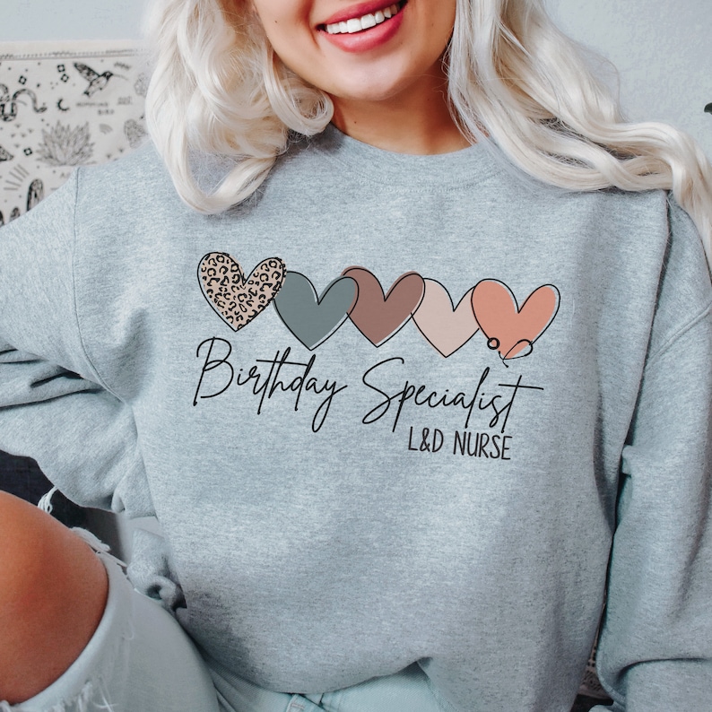 Op de afbeelding: Een lichtgrijs sweatshirt met een grafisch ontwerp van vijf harten op een rij. De harten zijn in tinten roze, bruin, blauw en luipaardprint. De tekst "Birthday Specialist L&D NURSE" staat onder de harten.