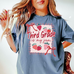 May include: Blue t-shirt with a strawberry-themed graphic. The graphic features the text "Third Grade is my jam" in red script, surrounded by strawberries and bows on a checkered background.