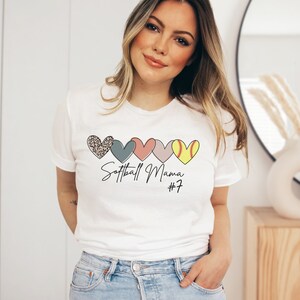 May include: White t-shirt with a graphic of five hearts in a row. The hearts are different colors and patterns, including leopard print, blue, pink, and yellow. The text "Softball Mama #7" is printed below the hearts.