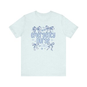 May include: Light blue t-shirt with a floral design and the text "emergency nurse" in blue. The design includes a bandage, a stethoscope, and a cross.