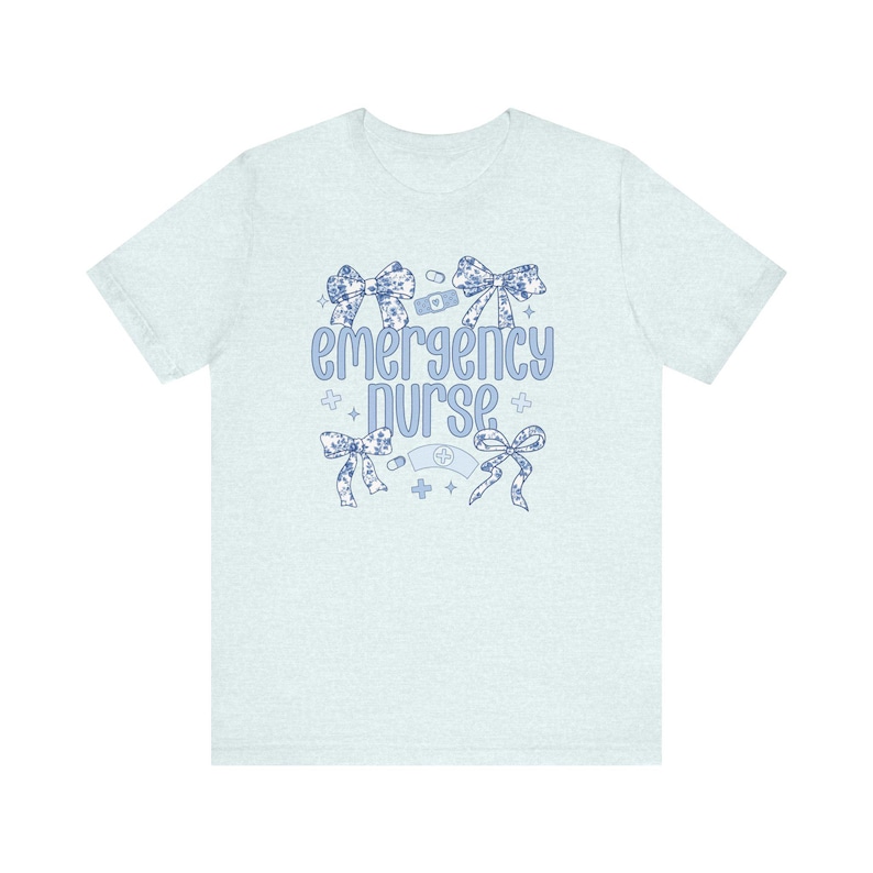 May include: Light blue t-shirt with a floral design and the text "emergency nurse" in blue. The design includes a bandage, a stethoscope, and a cross.