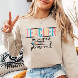 May include: A beige sweatshirt with the text "TEACHER in progress please wait" in colorful letters. The text is surrounded by a black line and a dashed line.