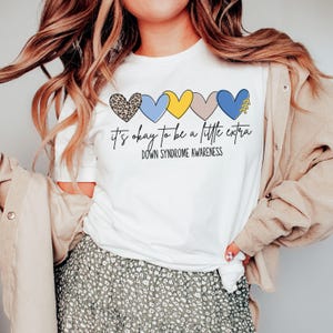 May include: White t-shirt with a row of colorful hearts and the text "it's okay to be a little extra" and "Down Syndrome Awareness".
