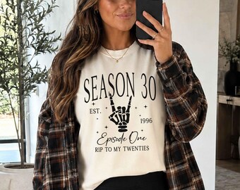 May include: A cream-colored crewneck sweatshirt featuring black text that reads SEASON 30, EST. 1996, Episode One, and RIP TO MY TWENTIES, accompanied by a graphic of a skeleton hand making a rock-and-roll gesture, worn under a brown plaid flannel shirt.