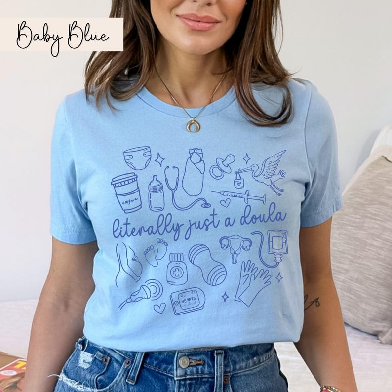 May include: Pale blue t-shirt featuring the phrase "literally just a doula" and line drawings of baby-related objects, such as a baby bottle and a stethoscope. The words "Baby Blue" are written in cursive at the top left.