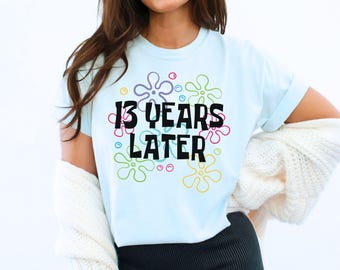 May include: Light blue t-shirt with a graphic of colorful cartoon flowers and the text "13 years later" in black.