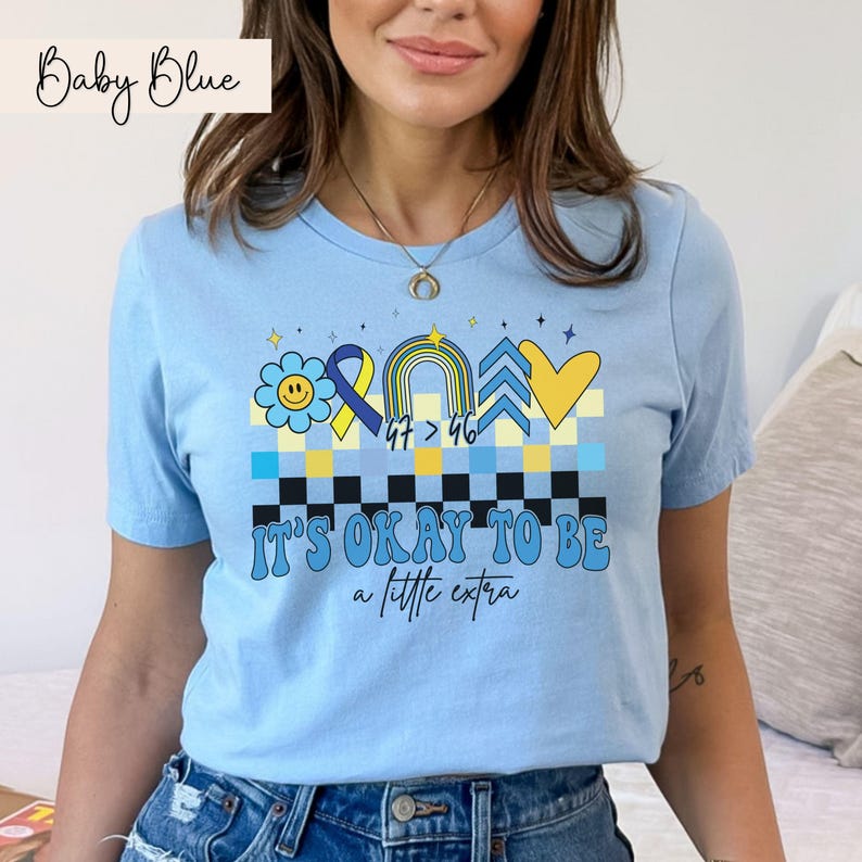 Pu&ograve; includere: Una maglietta blu chiaro con un design grafico che presenta un cuore giallo, frecce blu e gialle, un arcobaleno, un nastro blu, un fiore giallo con un viso sorridente e il testo "It's okay to be a little extra".