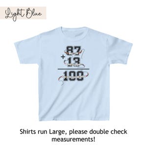 May include: Light blue t-shirt with a math equation printed on the front. The equation is 87 + 13 = 100. The numbers are made of colorful beads.