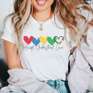 May include: White t-shirt with a row of colorful hearts, the last heart is a puzzle piece heart. The text below the hearts reads "Accept. Understand. Love."