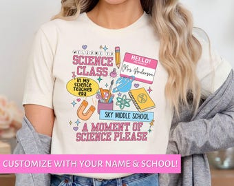 Pode incluir: Uma camiseta branca com um design colorido que apresenta ilustrações com tema científico, incluindo um béquer, um ímã, um caderno, um lápis e um planeta. O texto "Bem-vindo à aula de ciências" e "Um momento de ciência, por favor" também estão incluídos no design. O texto "Olá! Meu nome é Sra. Anderson" está em um crachá rosa.
