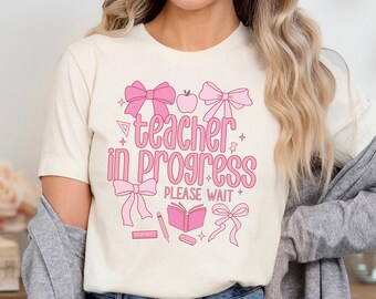 May include: A white t-shirt with a pink graphic that says "teacher in progress please wait". The graphic includes pink bows, an apple, a book, a pencil, a ruler, and an eraser.