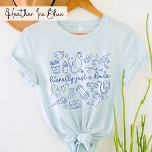 May include: Pale blue t-shirt featuring the phrase "literally just a doula" and blue illustrations of baby-related items. The shirt has rolled sleeves and is made of a soft material. The words "Heather Ice Blue" are at the top.