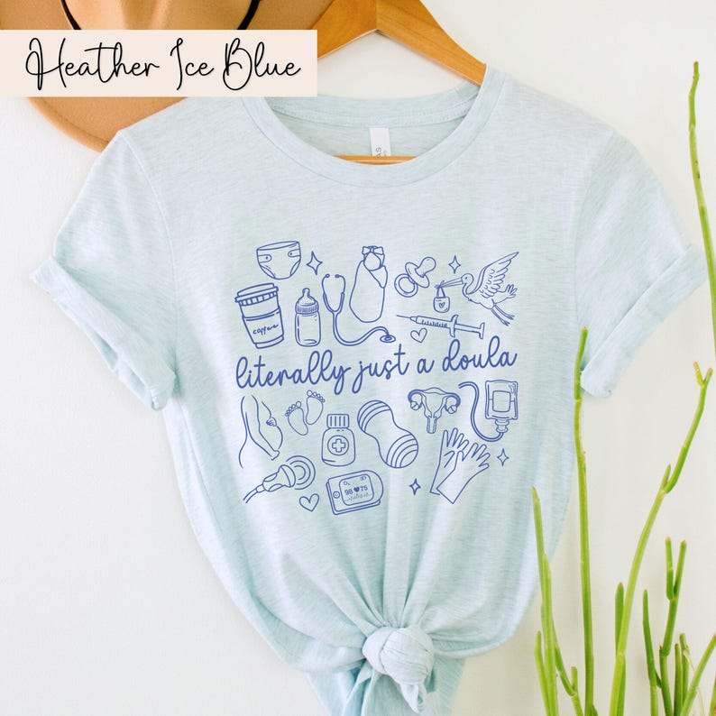 May include: Pale blue t-shirt featuring the phrase "literally just a doula" and blue illustrations of baby-related items. The shirt has rolled sleeves and is made of a soft material. The words "Heather Ice Blue" are at the top.