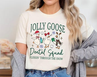 May include: Cream-colored t-shirt with the text "JOLLY GOOSE DENTAL SQUAD" in green. The shirt features three cartoon geese dressed in holiday attire, holding dental tools and floss. The phrase "FLOSSIN' THROUGH THE SNOW" is also printed on the shirt.