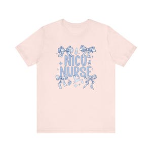 May include: Pink t-shirt with a blue design that says "NICU Nurse" surrounded by bows, baby bottles, and other baby-related imagery.