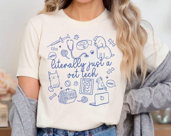 May include: A white t-shirt with a blue graphic design featuring a stethoscope, a bandage, a cat, a dog, a bird, a pet carrier, a ball, a laptop, and the text "literally just a vet tech".