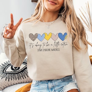 May include: A beige sweatshirt with a graphic of five colorful hearts and the text "it's okay to be a little extra Down Syndrome Awareness".
