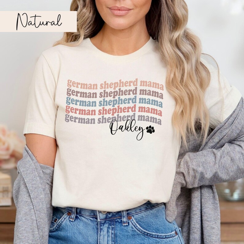 May include: A white t-shirt with the text "german shepherd mama" repeated five times in different colors. The text "Oakley" is written below the repeated text with a paw print.