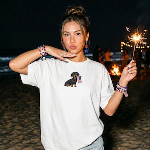 Op de afbeelding: Wit T-shirt met een afbeelding van een teckel die een Amerikaanse vlag vasthoudt. De hond is zwart en bruin. De persoon draagt armbanden en oorbellen met rode, witte en blauwe sterren. Een brandende sterretjes is in de hand van de persoon.